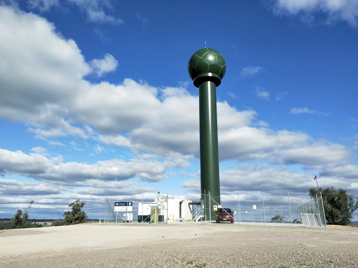 A forest-green radar tower with a large ball on top juts into a partly cloudy sky.